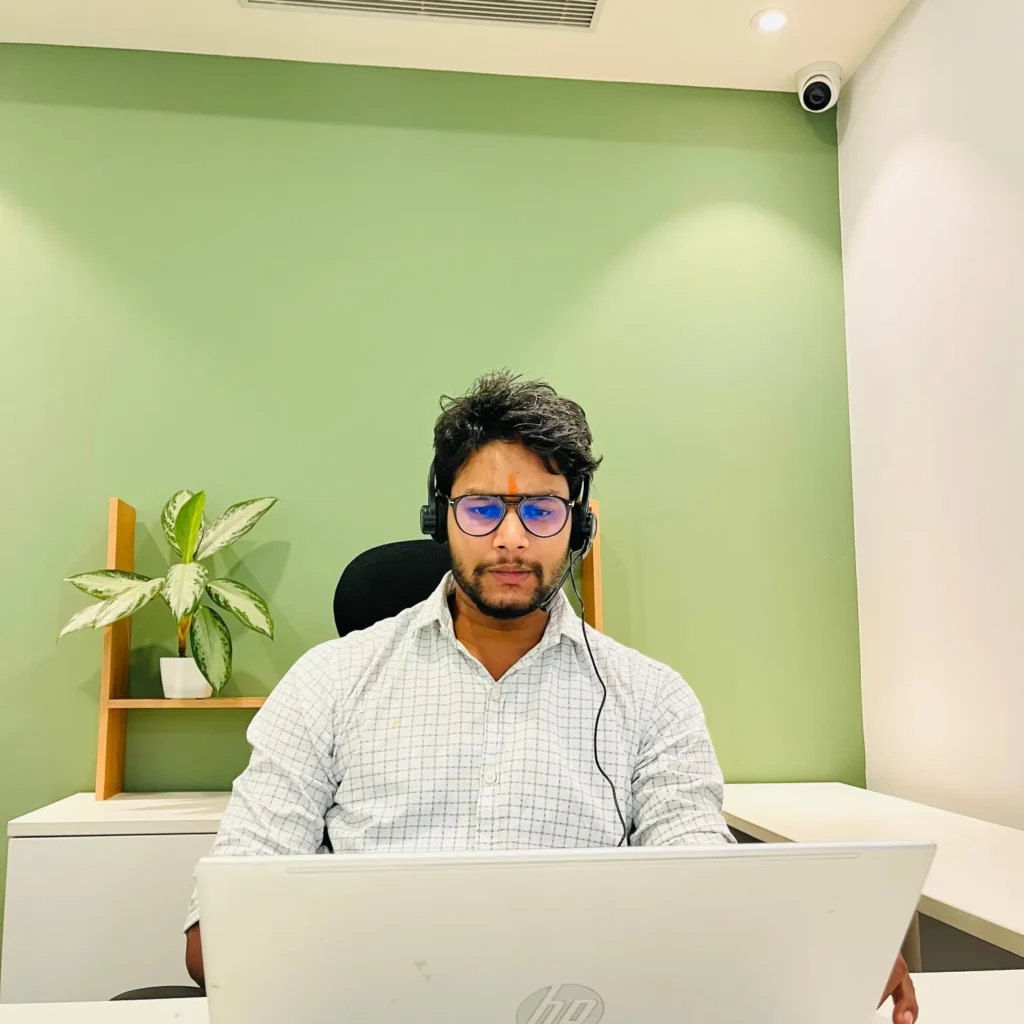 Portrait of Pravindra Yadav, founder of DML Training, sitting at a desk with a laptop and headphones, in an office setting with a green wall and plant.